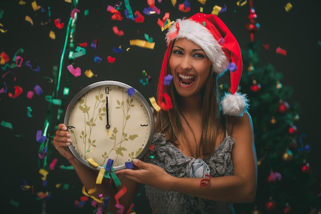 Foto hermosa mujer alegre celebrando el año nuevo y mostrando la medianoche en el reloj. ella se está divirtiendo, el confeti es el aire. mirando a la cámara.