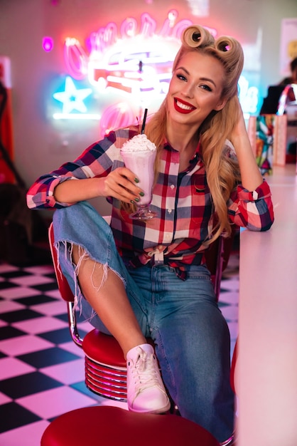 Hermosa mujer alegre con hermoso peinado bebiendo batido y sonriendo en retro american cafe