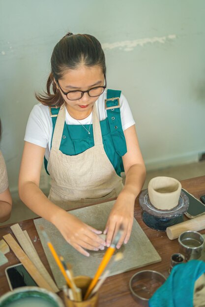 Hermosa mujer alfarera trabajando en la rueda de alfareros haciendo vasijas de cerámica de arcilla en el taller de cerámica Enfoque mano mujer joven adjuntando parte del producto de arcilla al futuro producto de cerámica Taller de cerámica