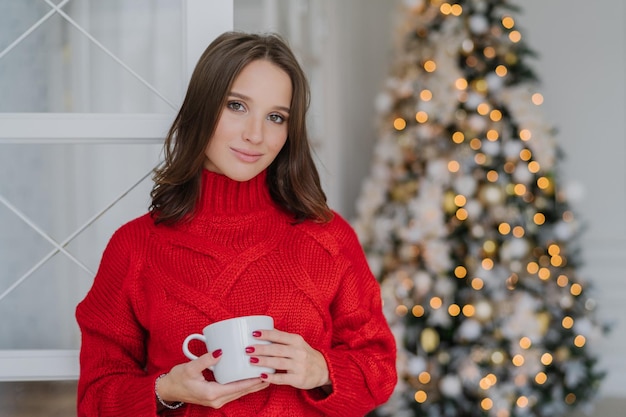 Hermosa mujer con apariencia agradable y maquillaje usa un cálido suéter de invierno de punto sostiene una taza de puestos de café cerca del árbol decorado de Año Nuevo planea cómo celebrar el concepto de descanso festivo