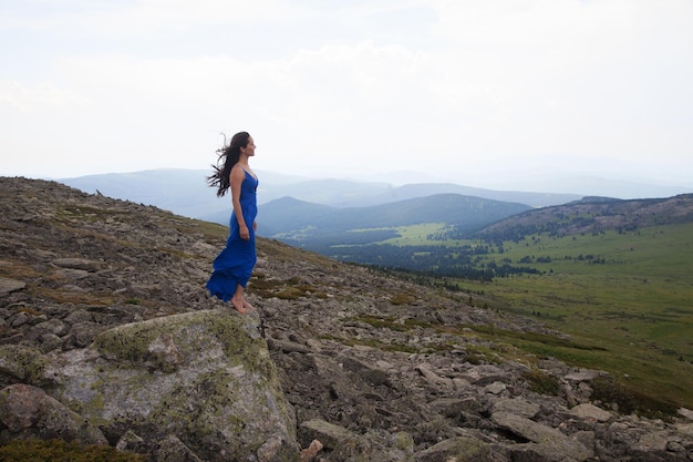 Una hermosa mujer de apariencia asiática con un vestido azul se encuentra en el borde de un acantilado entre las piedras en las montañas de Altai en verano. concepto de belleza, turismo.