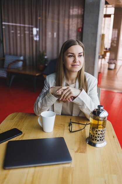 Una hermosa mujer de apariencia eslovena, una gerente con una chaqueta ligera y anteojos, una chica con una sonrisa en la cara, se sienta en una mesa de madera en un café. Trabajo remoto.