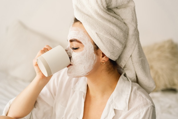 Hermosa mujer aplicando mascarilla facial y bebiendo té en la cama.