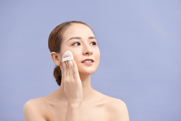 Hermosa mujer aplicando polvo seco con cojín cosmético en la piel de la cara. Foto de chica atractiva con maquillaje perfecto en pared beige. Concepto de belleza