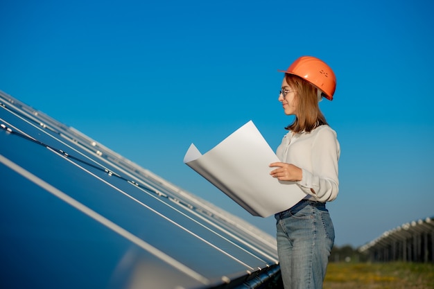 Hermosa mujer de arquitecto examinando un borrador de mapa o un plan de proyecto, actividad del trabajador mirando hacia la granja de células fotovoltaicas o campo de paneles solares.