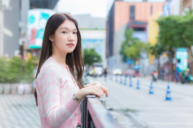 Hermosa mujer asiática con un abrigo de rayas rosas y blancas está de pie al aire libre urbano con ciudad y carretera