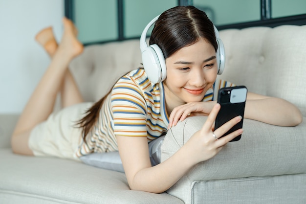 Hermosa mujer asiática acostada en el sofá y escuchando música mientras usa el teléfono
