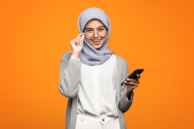 Hermosa mujer asiática alegre sosteniendo un teléfono inteligente en la pared naranja