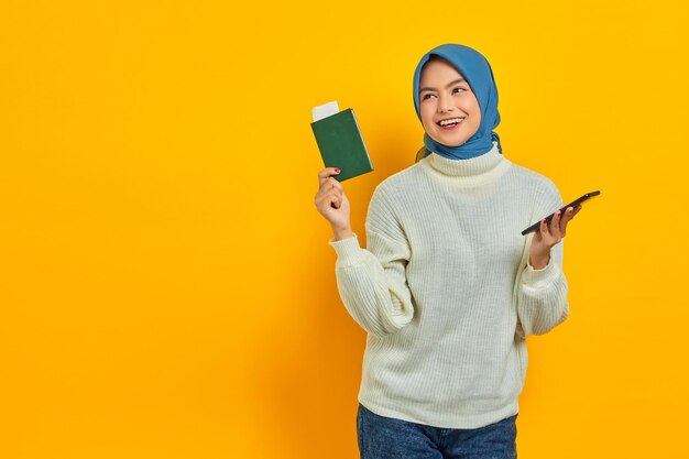 Hermosa mujer asiática alegre con suéter blanco sosteniendo teléfono móvil y pasaporte mirando a un lado el espacio de copia aislado sobre fondo amarillo Viaje de verano