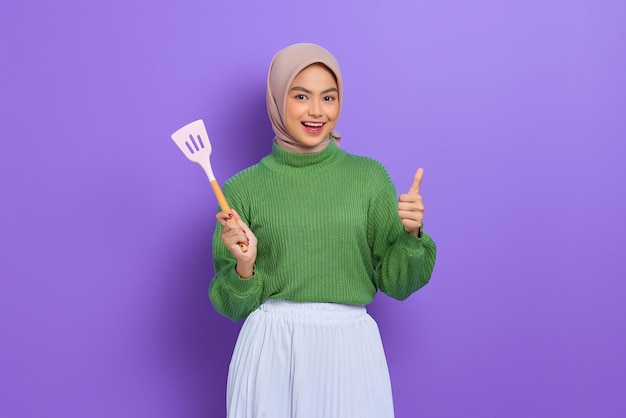 Hermosa mujer asiática alegre con suéter verde e hiyab sosteniendo una espátula y mostrando el pulgar hacia arriba aislado sobre fondo púrpura