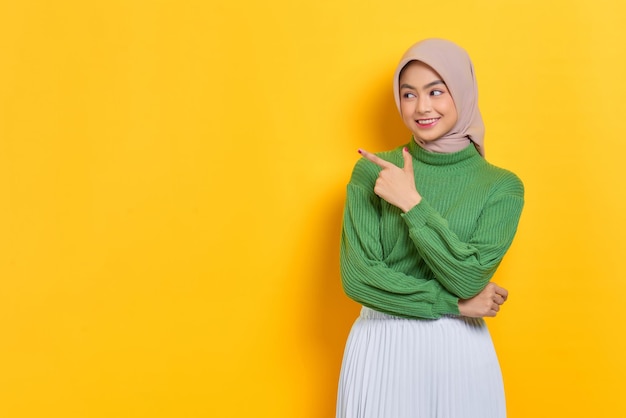 Hermosa mujer asiática alegre en suéter verde señalando con el dedo a un lado en el espacio de copia aislado sobre fondo amarillo