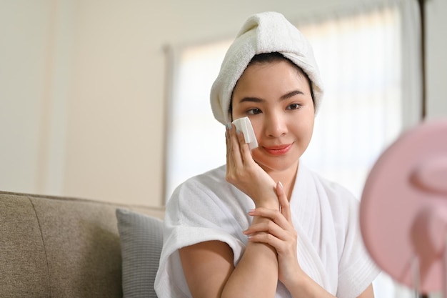 Hermosa mujer asiática en bata de baño limpiándose la cara haciendo tratamiento facial en casa