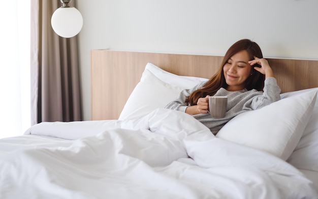 Una hermosa mujer asiática bebiendo café caliente mientras está acostado en una acogedora cama blanca en su casa por la mañana