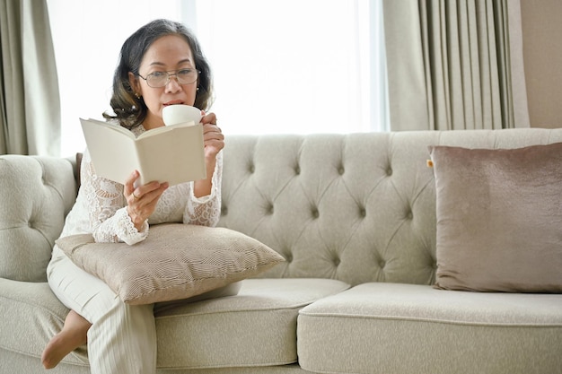 Hermosa mujer asiática bebiendo café matutino mientras lee un libro en su sala de estar