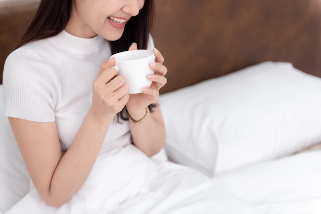 Hermosa mujer asiática bebiendo café y una sonrisa feliz en la cama en el dormitorio blanco