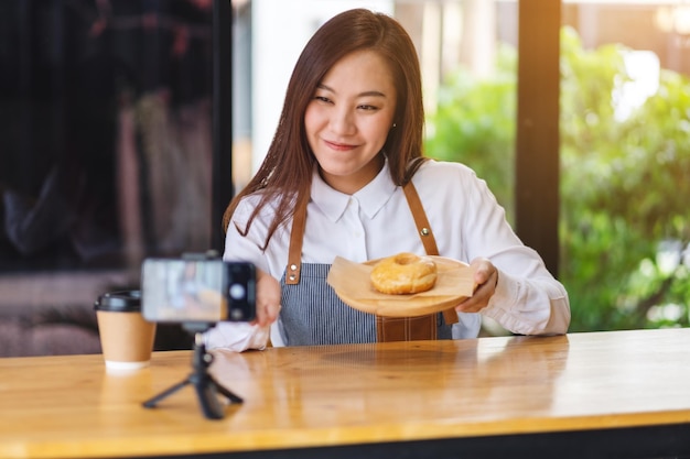 Una hermosa mujer asiática blogger de comida o vlogger que muestra un trozo de rosquilla mientras graba un video en la cámara