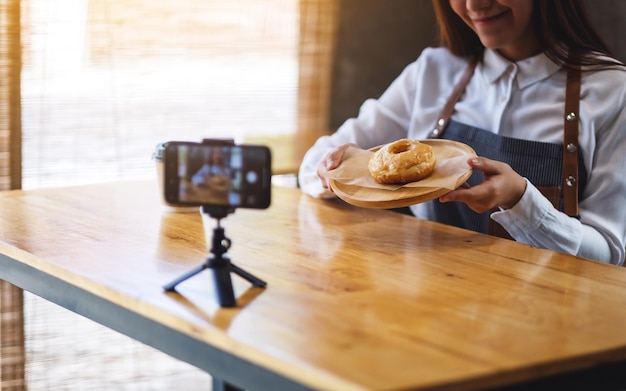 Una hermosa mujer asiática blogger de comida o vlogger que muestra un trozo de rosquilla mientras graba un video en la cámara