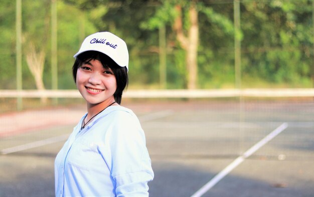 Hermosa mujer asiática con cabello corto, sombrero y una amplia sonrisa en la cancha de tenis