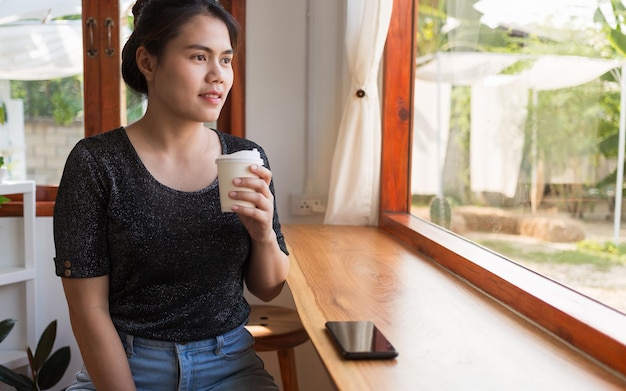 Hermosa mujer asiática en una cafetería sosteniendo una taza de café de papel sonriendo y relajada en un café