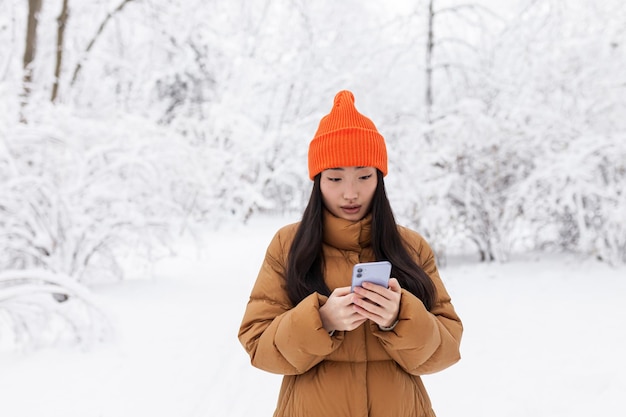 Hermosa mujer asiática caminando en el parque, usa el teléfono para comprar en línea, en un día nevado de invierno