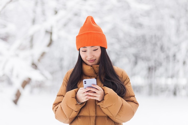 Hermosa mujer asiática caminando en el parque, usa el teléfono para comprar en línea, en un día nevado de invierno