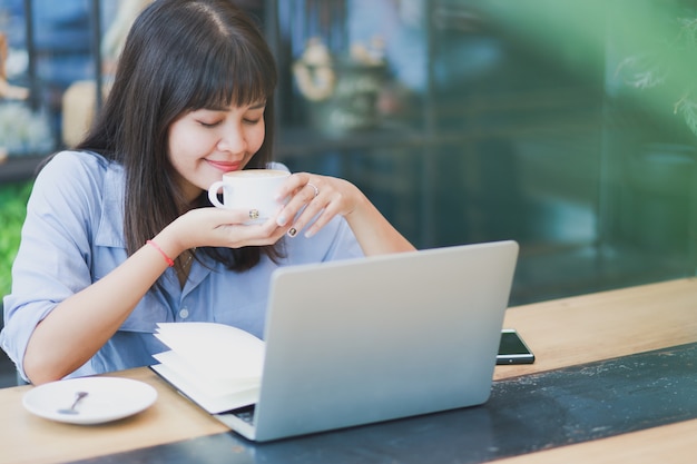 Hermosa mujer asiática en camisa azul usando laptop y tomando café