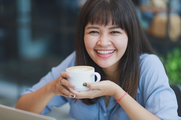 Hermosa mujer asiática en camisa azul usando laptop y tomando café