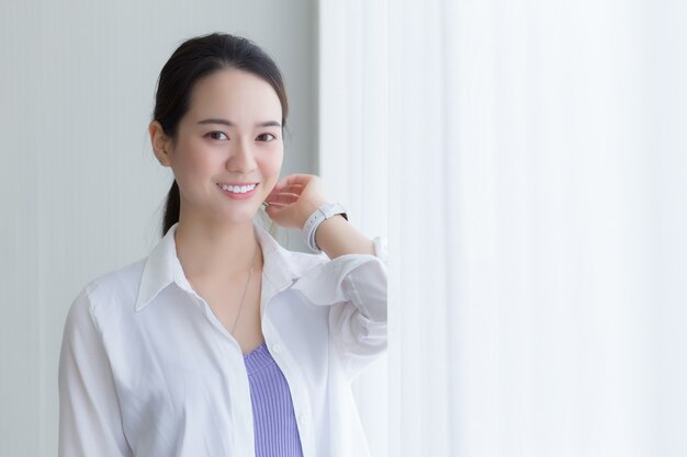 Hermosa mujer asiática en camisa blanca está sonriendo y de pie junto a la ventana con cortina blanca.