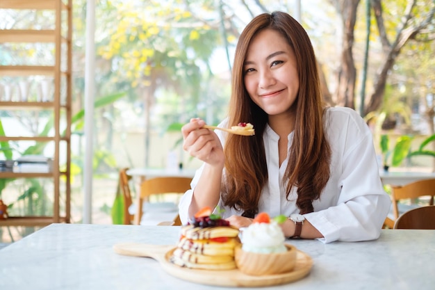 Una hermosa mujer asiática comiendo panqueques de bayas mixtas con helado y crema batida con cuchara de madera