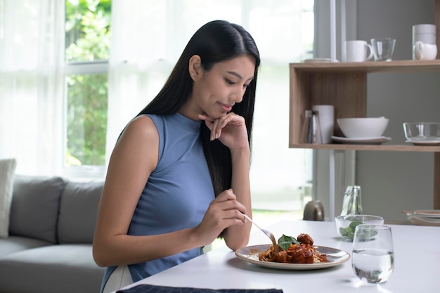 Hermosa mujer asiática comiendo pasta en la mesa por la mañana en casa Sonriente mujer sana comiendo espaguetis mientras se sienta y desayuna en la mesa de la cocina