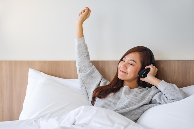 Una hermosa mujer asiática disfruta escuchando música con auriculares mientras está acostada en una acogedora cama blanca en casa