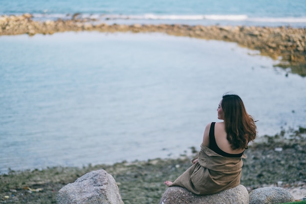 Foto una hermosa mujer asiática disfruta sentada en la roca junto a la orilla del mar