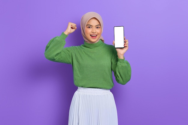 Hermosa mujer asiática emocionada con suéter verde e hiyab mostrando un teléfono móvil con pantalla en blanco aislado sobre un fondo morado