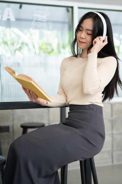 Una hermosa mujer asiática está escuchando música y leyendo un libro mientras se relaja en una cafetería
