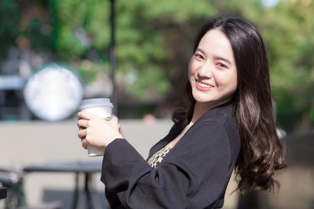 Hermosa mujer asiática está sentada en una silla y sonriendo felizmente frente a la cafetería.