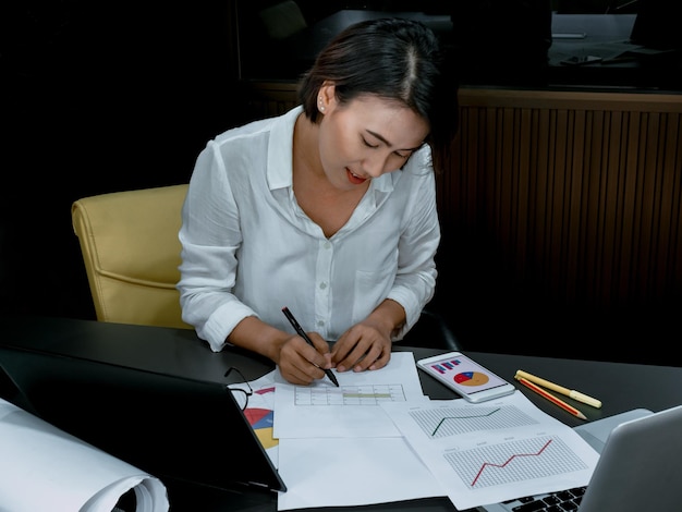 Hermosa mujer asiática feliz trabajando horas extras con una computadora portátil en el escritorio de la oficina