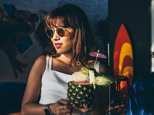Hermosa mujer asiática feliz vestida de blanco sin mangas y gafas de sol sosteniendo un cóctel de jugo de piña en verano
