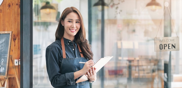 Hermosa mujer asiática joven barista en delantal sosteniendo cllpboard y de pie frente a la puerta del café con letrero abierto. Concepto de inicio de propietario de negocio.