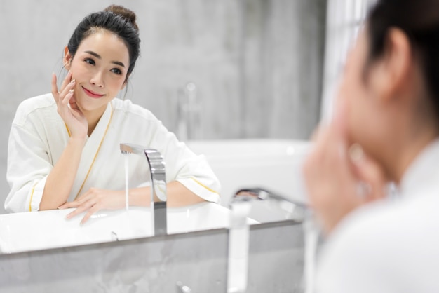 Hermosa mujer asiática joven que se lava la cara limpia con agua y sonriendo frente al espejo en el baño. Belleza y spa. Piel fresca perfecta