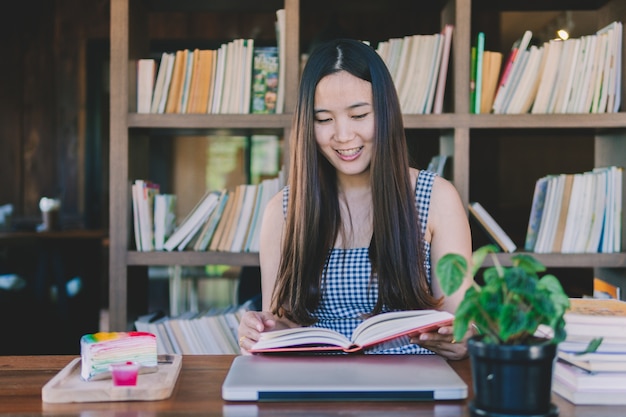 Hermosa mujer asiática joven sentado y leyendo el libro en la biblioteca con estante de libros en centrico