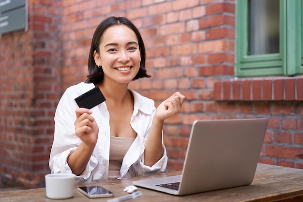Hermosa mujer asiática joven con tarjeta de crédito sentada cerca de la computadora portátil y sonriendo pagando facturas tiendas onli