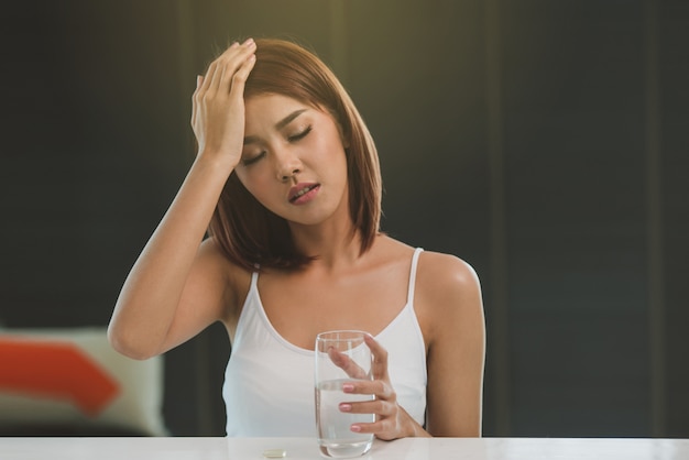 Hermosa mujer asiática joven tomando píldora contra dolor de cabeza en casa