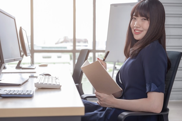 Una hermosa mujer asiática lleva un traje azul oscuro sentada en un escritorio en una oficina moderna y está feliz de trabajar y tiene un gran fondo de ventana de vidrio.