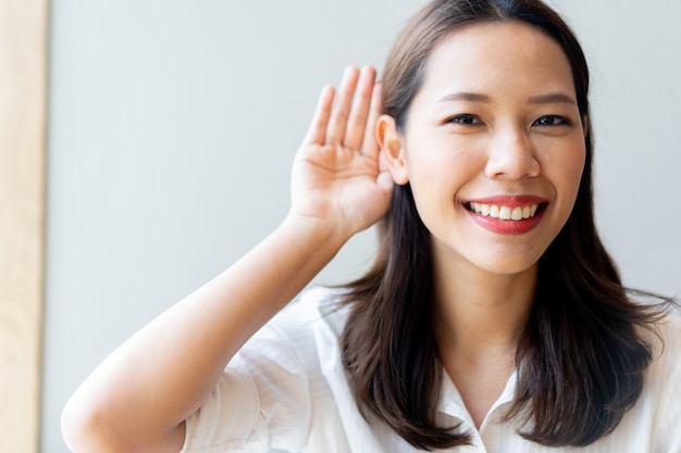 Hermosa mujer asiática médico sonriendo y usar la mano detrás de la oreja para tratar de escuchar el concepto de sordera