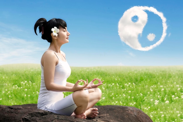 Hermosa mujer asiática meditando al aire libre en la roca en el prado con el símbolo de nubes yin yang en el cielo