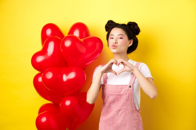 Hermosa mujer asiática mostrando corazón, te amo gesto y besos labios, de pie cerca de románticos globos rojos. Concepto de día de San Valentín.