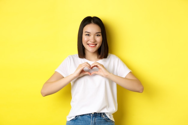 Hermosa mujer asiática mostrando te amo gesto de corazón, sonriendo a la cámara, de pie contra el fondo amarillo. Concepto de día de San Valentín y romance.