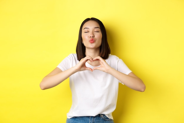 Hermosa mujer asiática mostrando te amo gesto de corazón, sonriendo a la cámara, de pie contra el fondo amarillo. Concepto de día de San Valentín y romance.