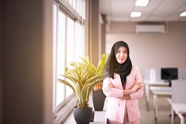 Hermosa mujer asiática de negocios con hijab mientras está de pie y mirando por la ventana en la oficina