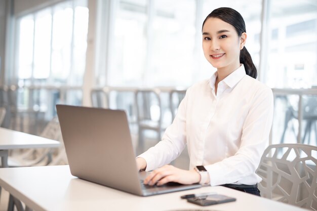 Hermosa mujer asiática de negocios con traje trabajando en un escritorio de oficina usando una computadora con un teléfono inteligente.